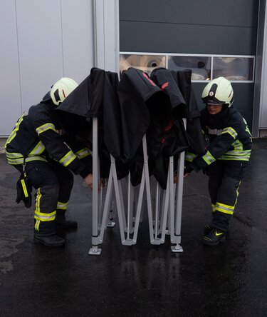 Firefighters from Ulm, Germany opening a custom black 3x3 m Ecotent pop-up gazebo with red lettering.