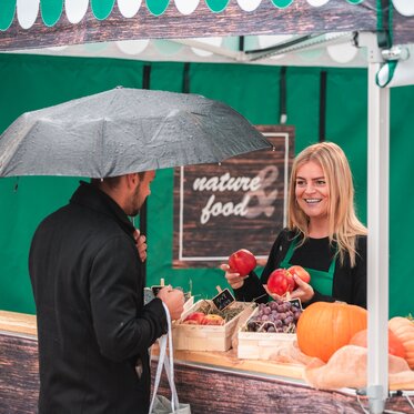 The saleswoman in the market tent holds various fruits in her hand. The customer is standing in front of the sales tent at the counter.