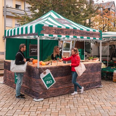 The market tent is fully printed and is used at the weekly market and farmers' market. 2 customers look at the products.