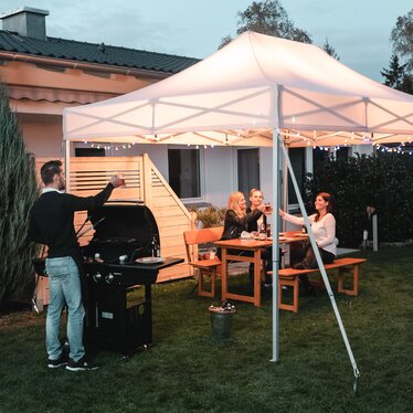 Friends celebrate a barbecue and garden party under the garden tent. The garden tent is festively decorated with fairy lights.