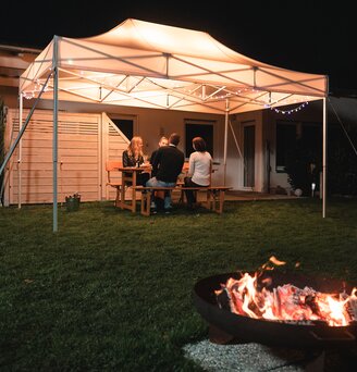The 4.5x3 m garden tent stands in the garden in the dark. The 4 guests sit under it. There is a fireplace in front of the gazebo.